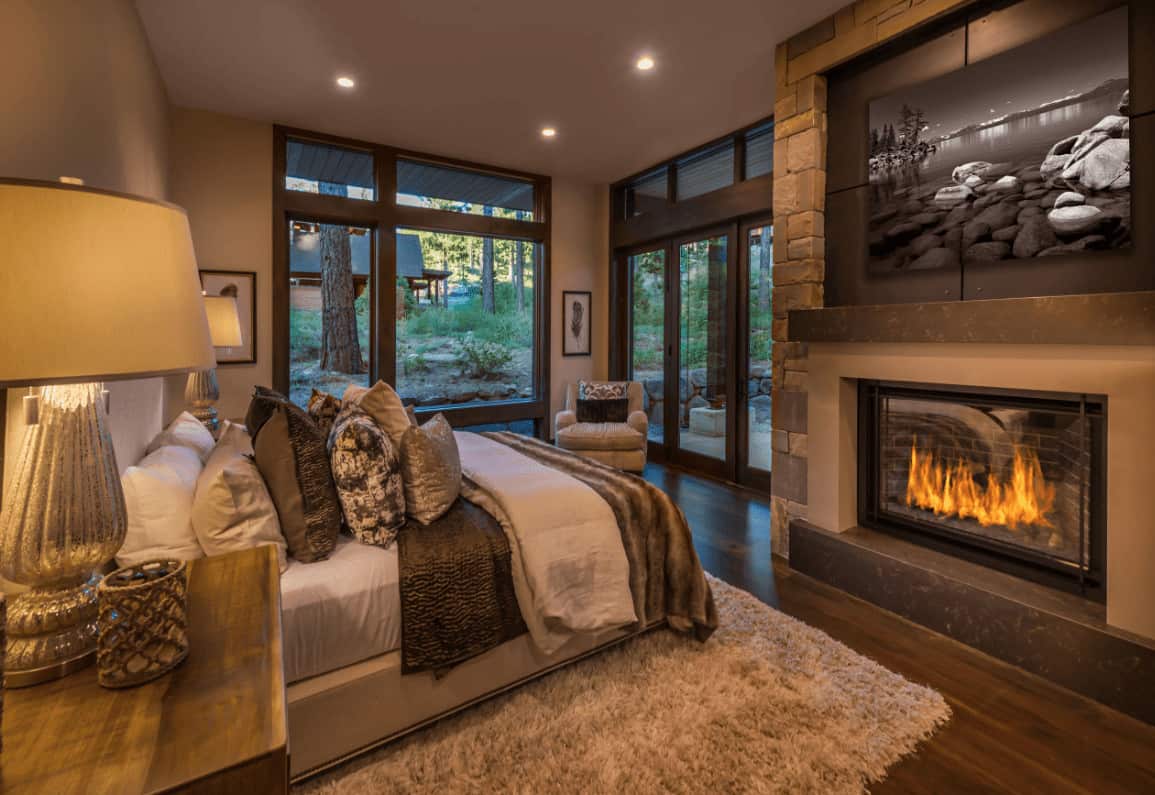 The warm primary bedroom boasts a comfy bed and a fireplace with a landscape canvas on top. It has full height glazing and hardwood flooring topped by a gray shaggy rug.
