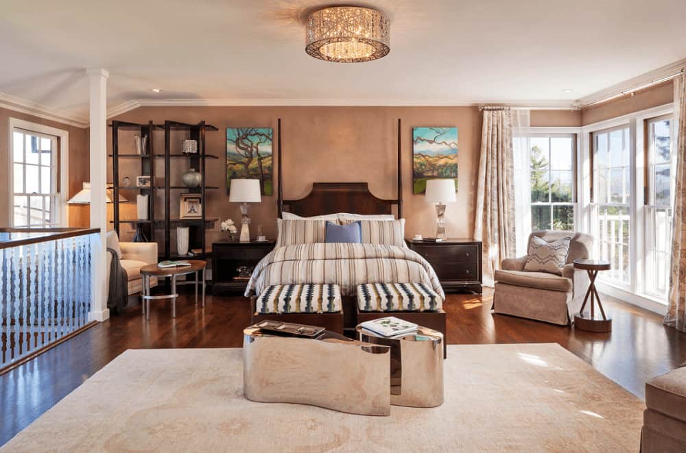This primary bedroom is decorated with a fancy flush mount light and lovely tree wall arts that hung over the dark wood nightstands. It has beige seats and a wooden bed with striped ottoman and stylish mirrored tables on its end.
