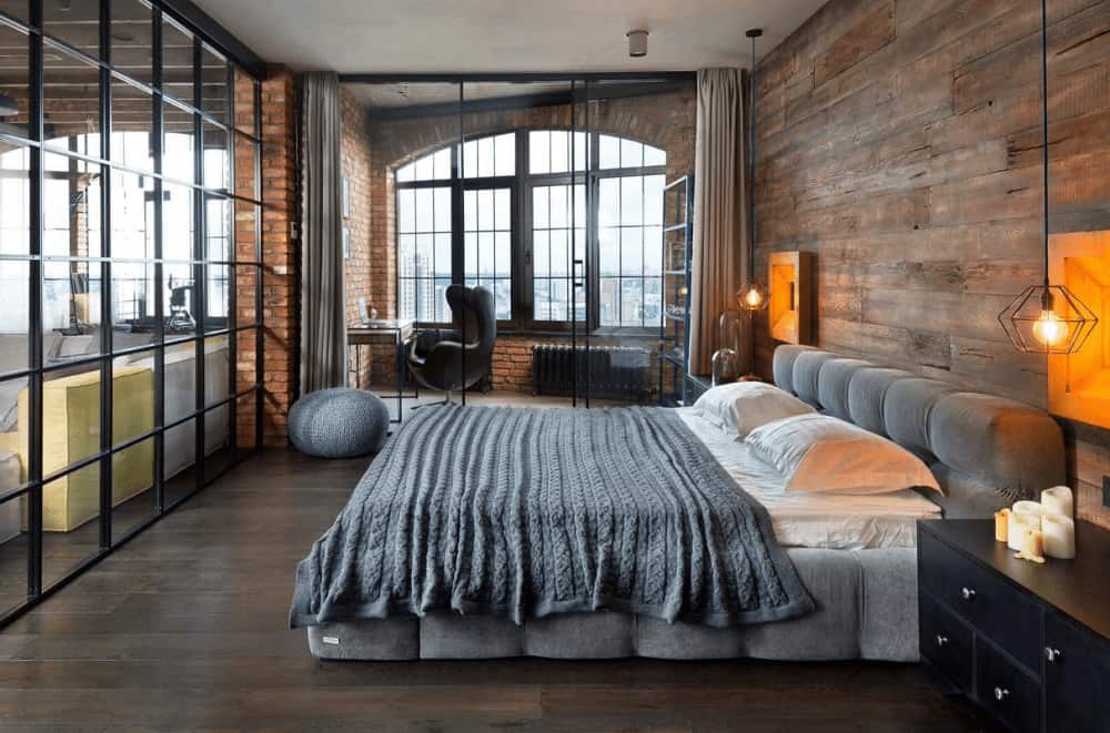 Industrial primary bedroom illuminated by geometric pendant lights that hung over the black nightstands flanking a gray tufted bed. It has wide plank flooring and a glazed door that leads to the office area.
