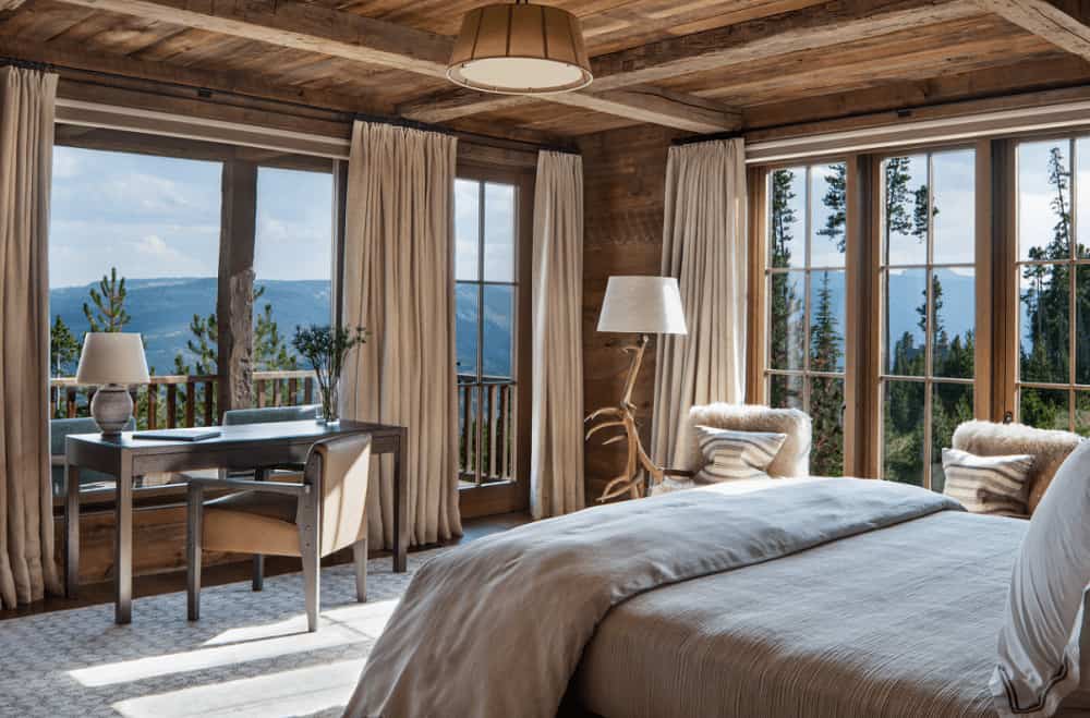 Brown primary bedroom surrounded with floor to ceiling windows that overlook the stunning mountain. It has a wooden desk and a cozy bed illuminated by a pendant light.