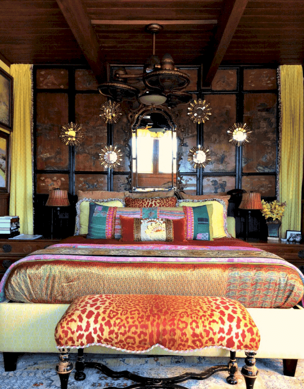 An animal printed bench sits in front of the yellow patterned bed decorated with sunburst and ornate arched mirrors. It is lighted by a unique chandelier that hung from the wood beam ceiling.