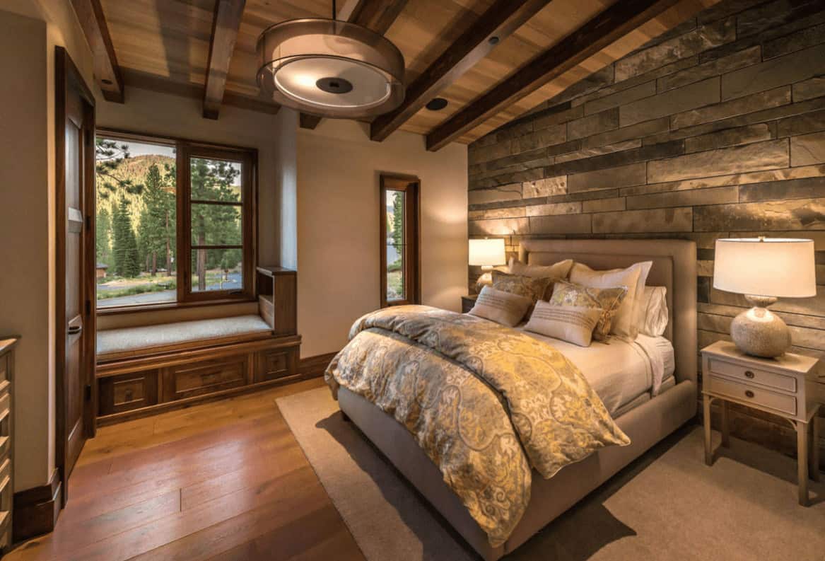A stone accent wall adds texture in this primary bedroom with a window seat nook and beige upholstered bed lighted by a pair of table lamps along with a drum pendant light.