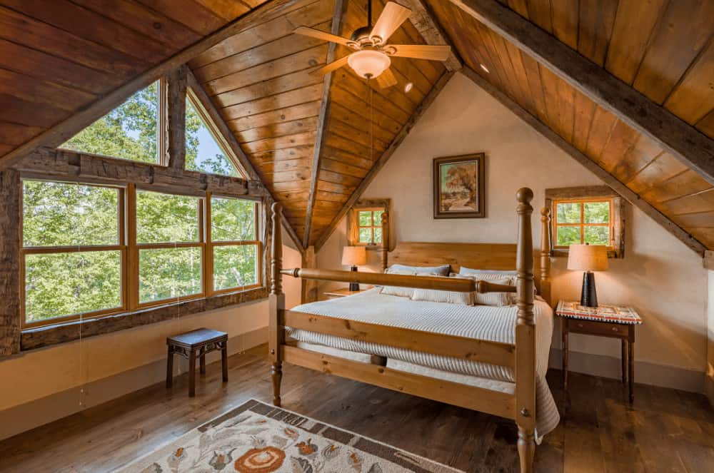 A framed wall art hangs above the wooden bed in this rustic bedroom with glass paneled windows and vaulted ceiling clad in natural wood planks.
