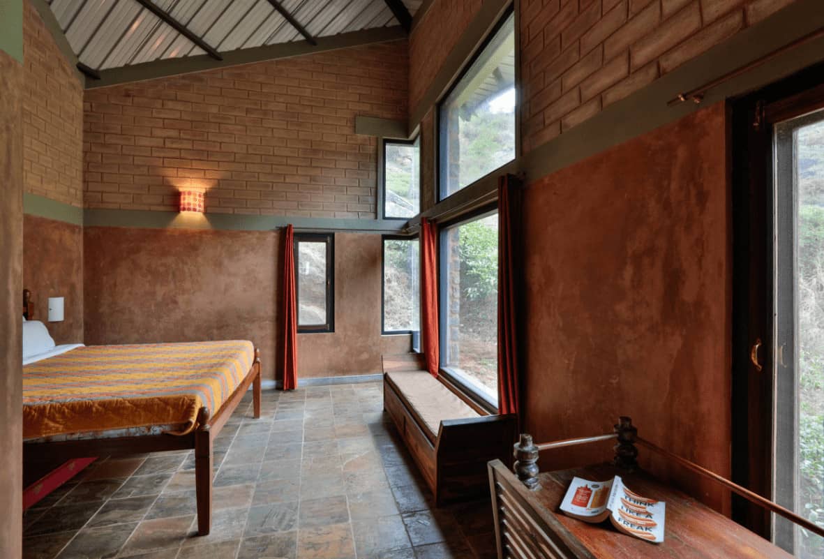Brown bedroom with a wooden bed and dotted sconce mounted on the upper brick wall. It has limestone flooring and picture windows that allow natural light in.