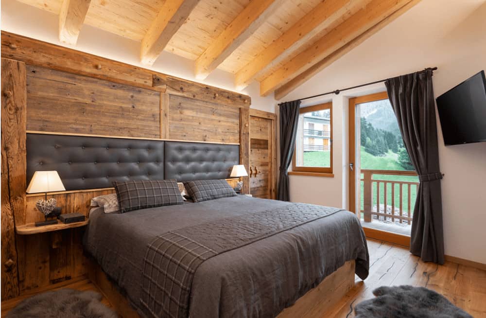 This primary bedroom showcases a wall mount TV and a wooden bed with custom tufted headboards fitted on the wood paneled wall. It includes gray drapes and shaggy rugs that lay on the hardwood flooring.