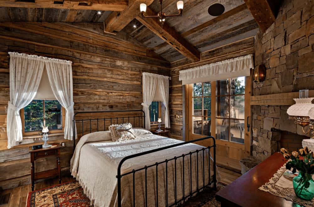 Rustic bedroom with a black metal bed and wooden nightstand topped with a green translucent flower vase. It is illuminated by recessed lights and a brass chandelier that hung from the vaulted ceiling.