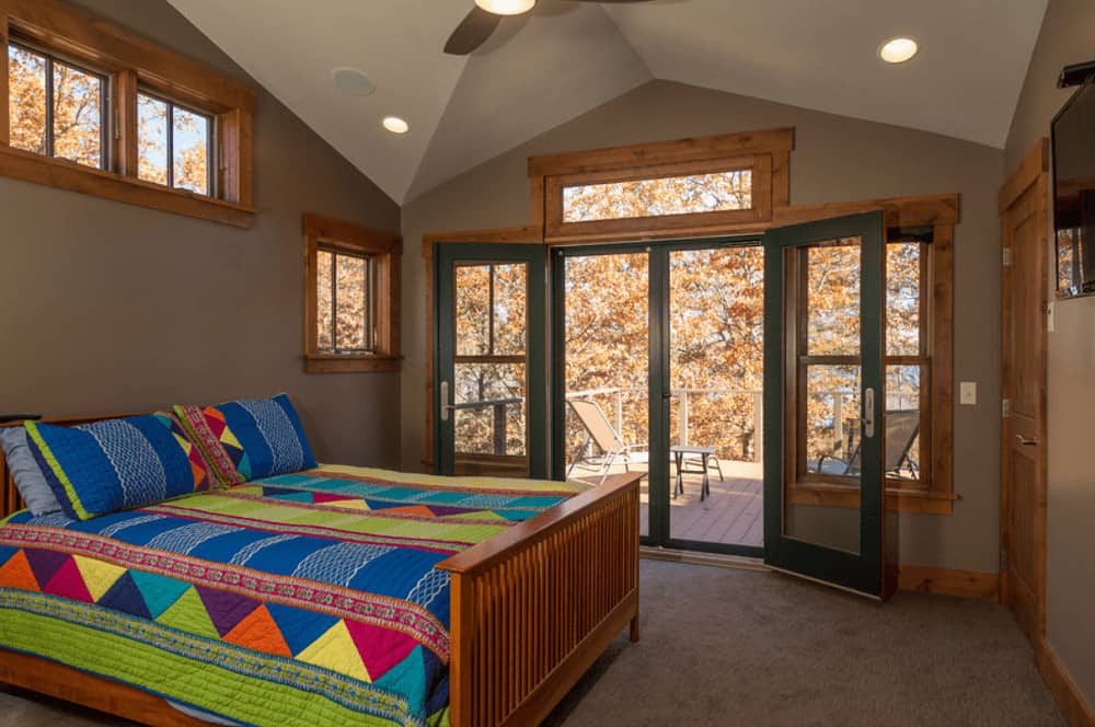 A wooden bed dressed in brightly multi-colored bedding stands out in this brown primary bedroom with carpet flooring and glass double door that leads out to the balcony.