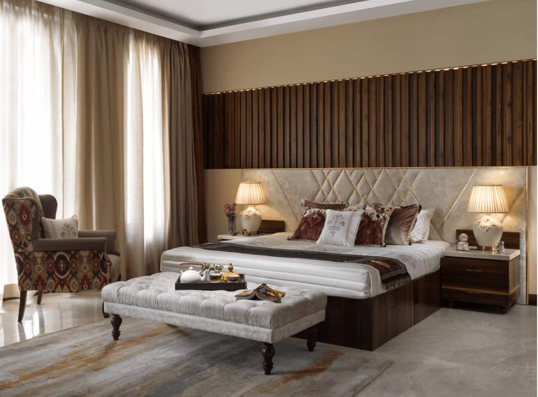 Victorian bedroom features a patterned wingback chair and a wooden bed with a classy custom headboard. It is accompanied by a gray tufted bench and wooden nightstands topped with white table lamps.