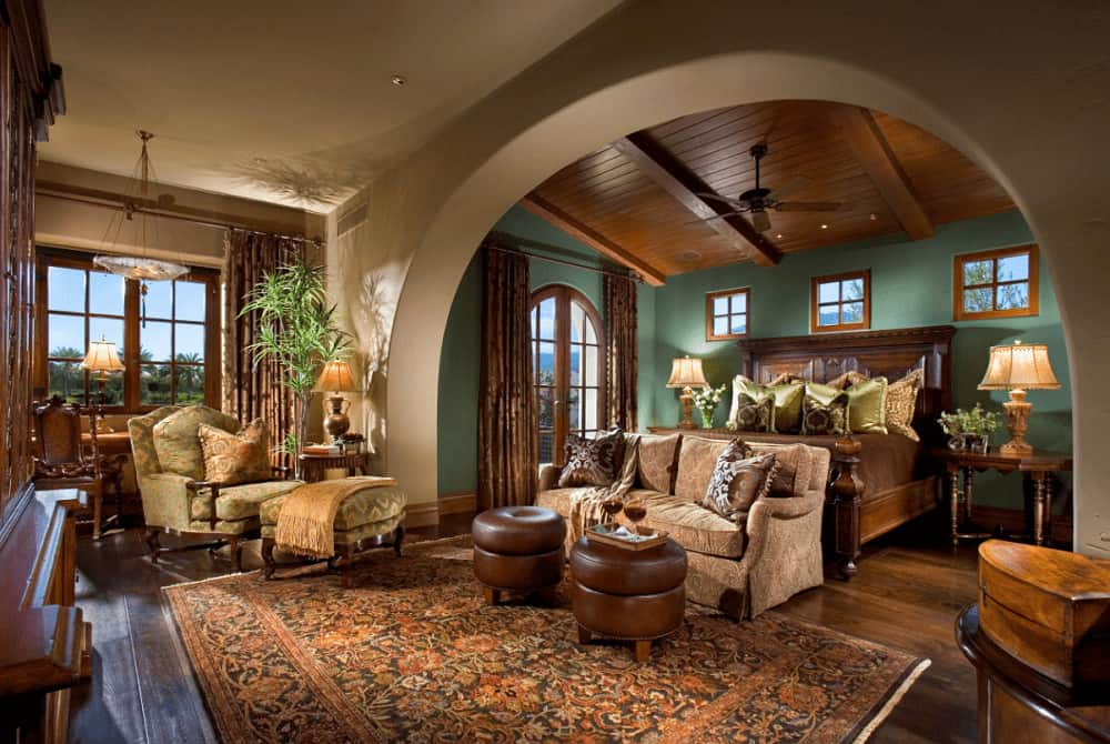Mediterranean bedroom with an open archway in the middle framing the wooden bed with a skirted sofa and brown leather ottomans on its end. It is accompanied by a green lounge chair and nightstands topped with lovely table lamps.