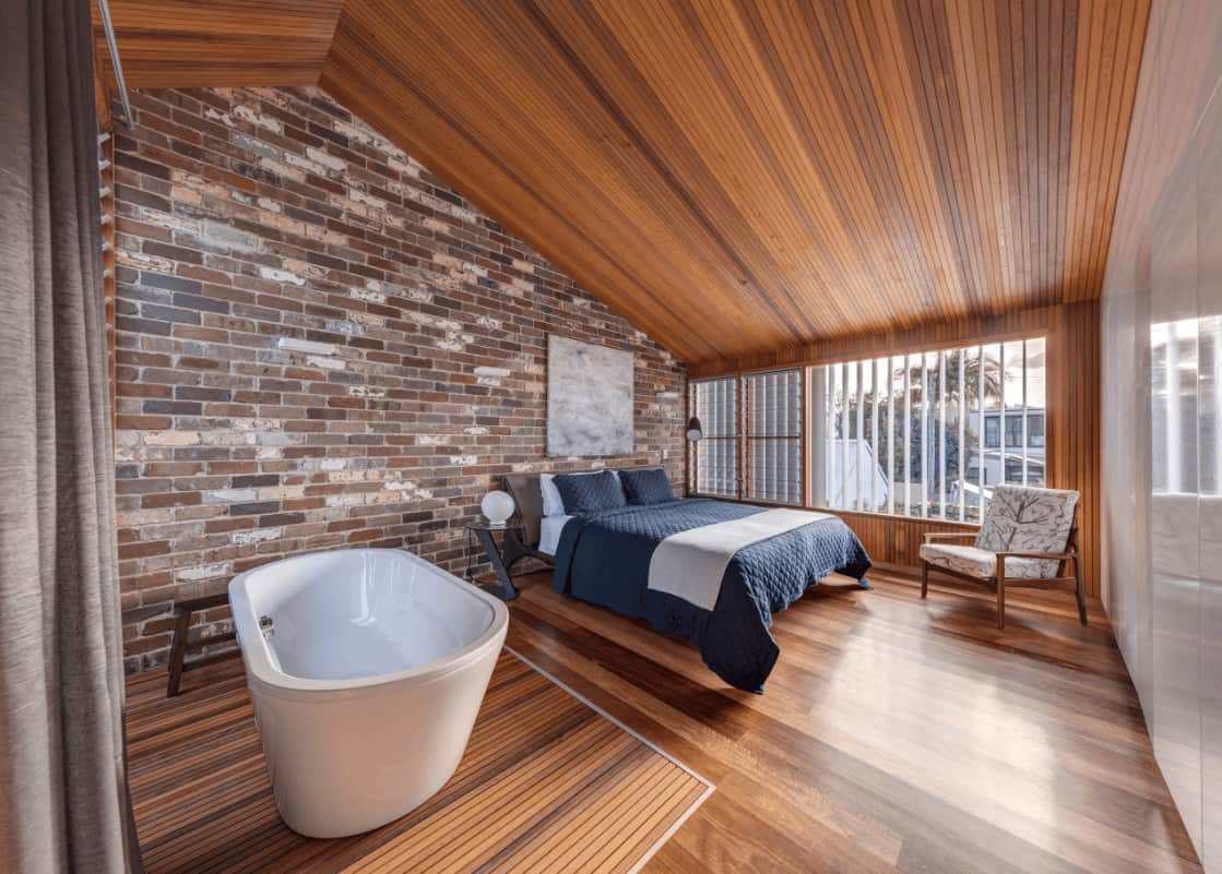 This primary bedroom boasts a cozy bed with a gray artwork on top mounted on the brick accent wall. It is accompanied by a printed armchair and a freestanding tub on the side.