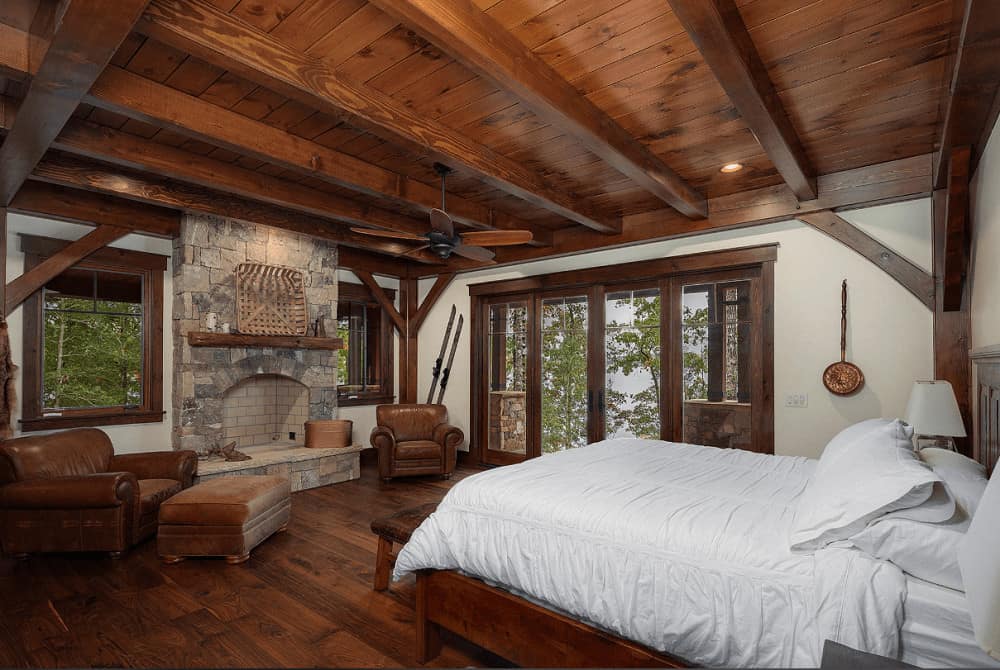 A wooden bed dressed in white bedding faces the stone brick fireplace topped with a rustic mantel and wicker wall art. It is flanked by brown leather armchairs over wide plank flooring.