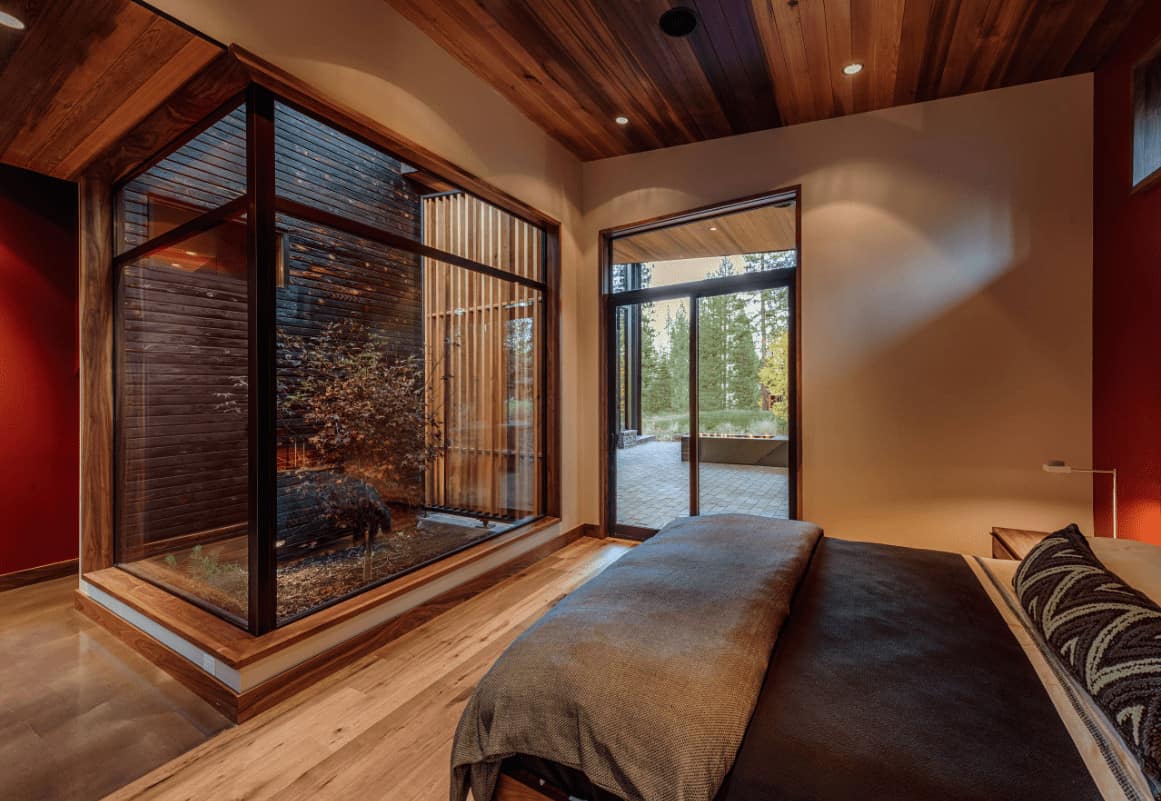 The sleek primary bedroom offers a comfy bed and a lovely plant enclosed in aluminum framed glass. It has light hardwood flooring and wood plank ceiling mounted with recessed lights.