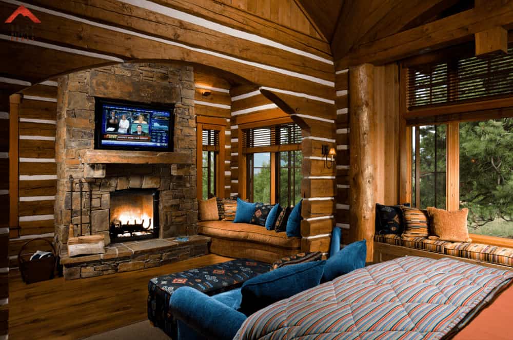 Blue velvet pillows and sofa add a nice accent in this brown primary bedroom with window seat nooks and wall mount TV that hung above the stone brick fireplace.
