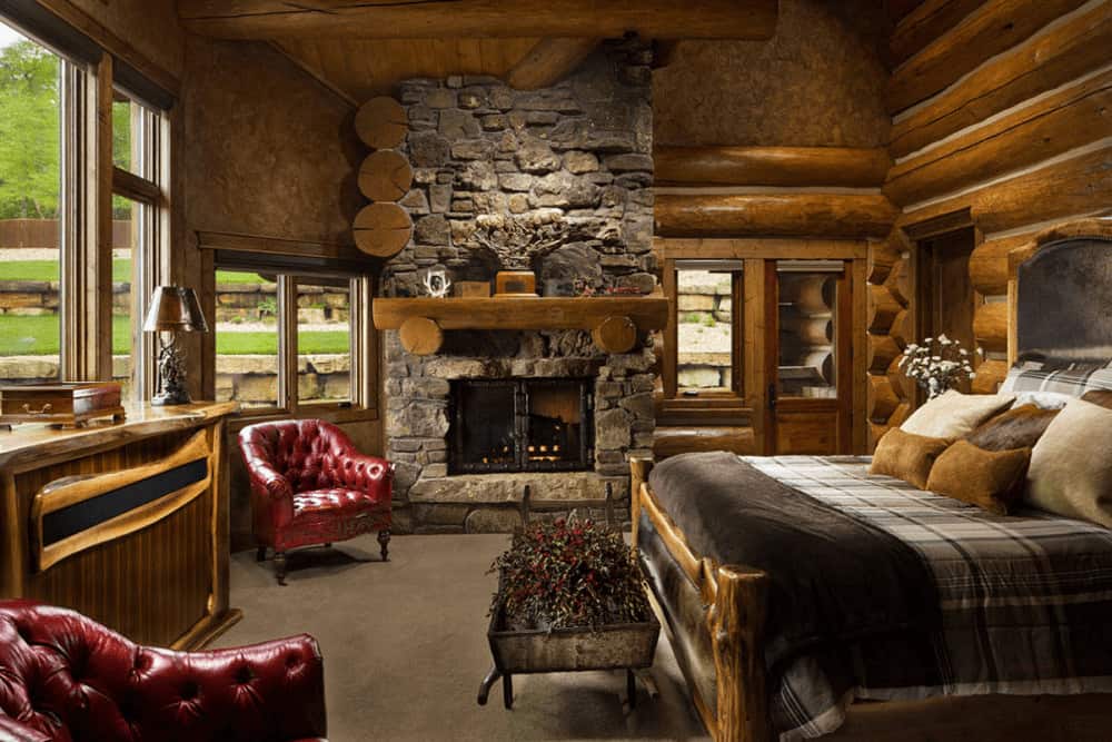 Fluffy earthy pillows lay on the cozy bed in this primary bedroom boasting red chesterfield chairs and a stone fireplace lined with a wooden mantel.