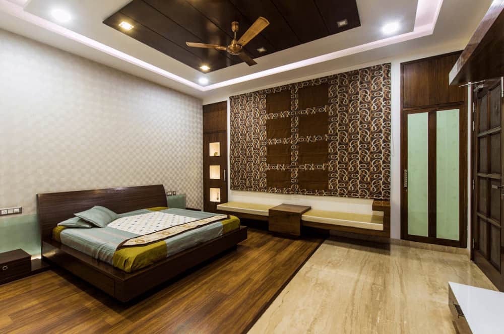This primary bedroom offers a platform bed and a sleek seat on the side accented by a large patterned tapestry. It has hardwood flooring and tray ceiling mounted with a bronze fan.