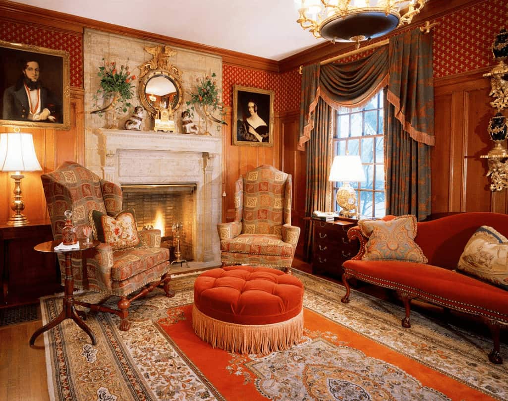 Elegant living room clad in red patterned wallpaper and wooden wainscoting mounted with lovely portraits. It has a fireplace and classy seats over a bordered area rug.