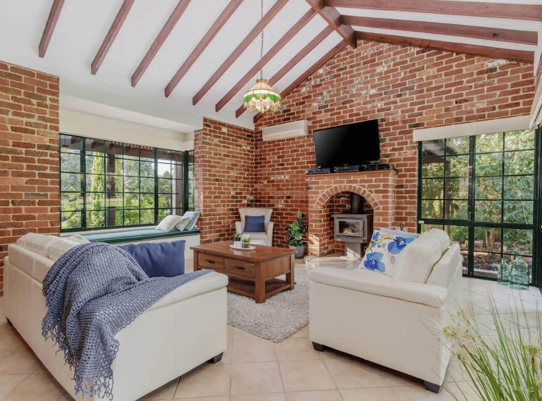 Airy living room with a window seat nook and cozy, white seats paired with a wooden coffee table that's lighted by a gorgeous pendant light. It includes a flat-screen TV and a fireplace fixed on the red brick walls.