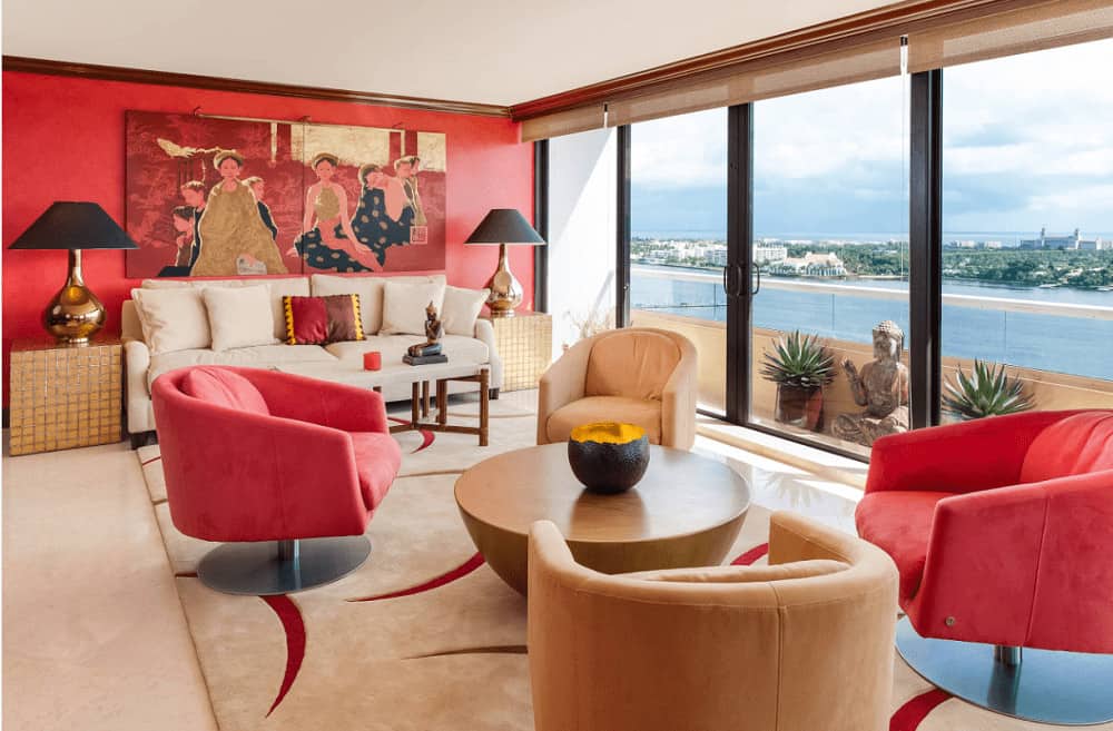 A multi-panel painting hangs over the beige sofa accompanied by cube end tables and sleek chairs surrounding a wooden coffee table. It has carpet flooring and full height glazing overlooking a spectacular lake view.