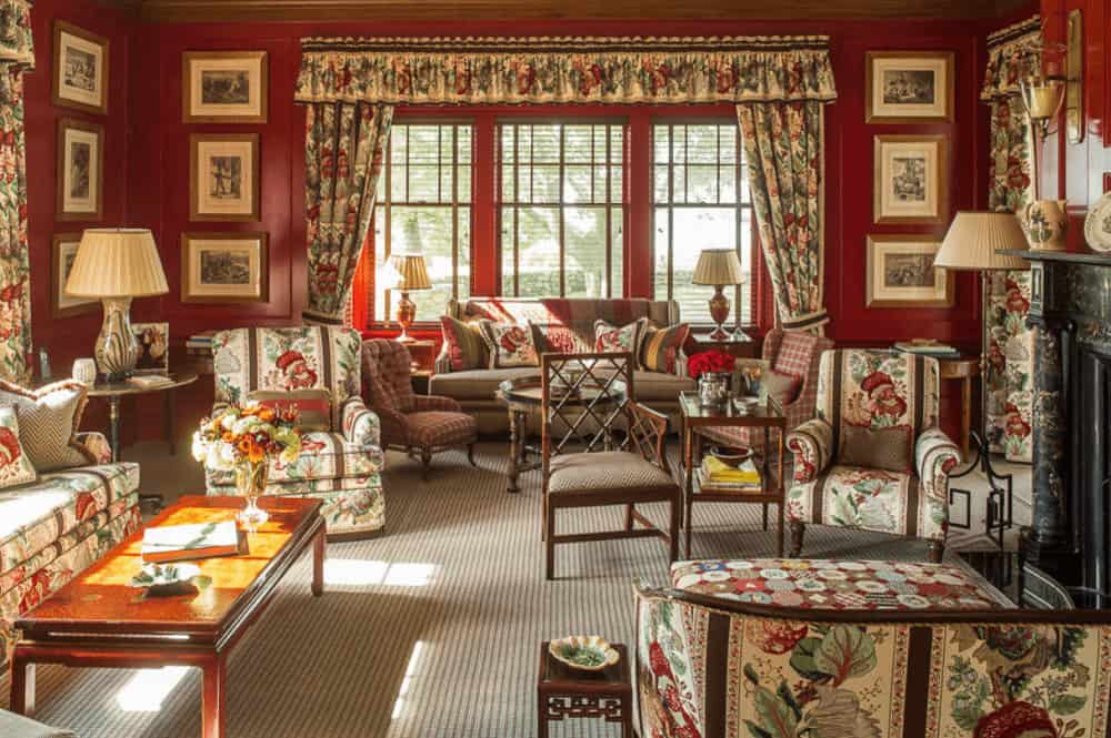 Floral draperies and valances complement the armchairs over a striped area rug. This room has wooden framed windows and red wainscoted walls filled with black and white photos.