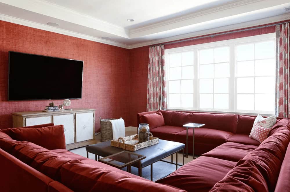 Natural light flows in through the white framed windows in this red living room with a sectional sofa and a leather coffee table topped with a wicker tray. It includes a wall mount TV that hung above the wooden stand.