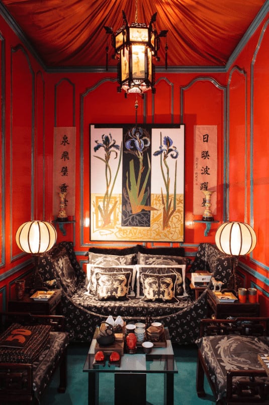 Small living room with classy sits and gorgeous artwork mounted on the red wainscoted walls. It is illuminated by round table lamps and a lantern pendant light that hung from the canopy ceiling.