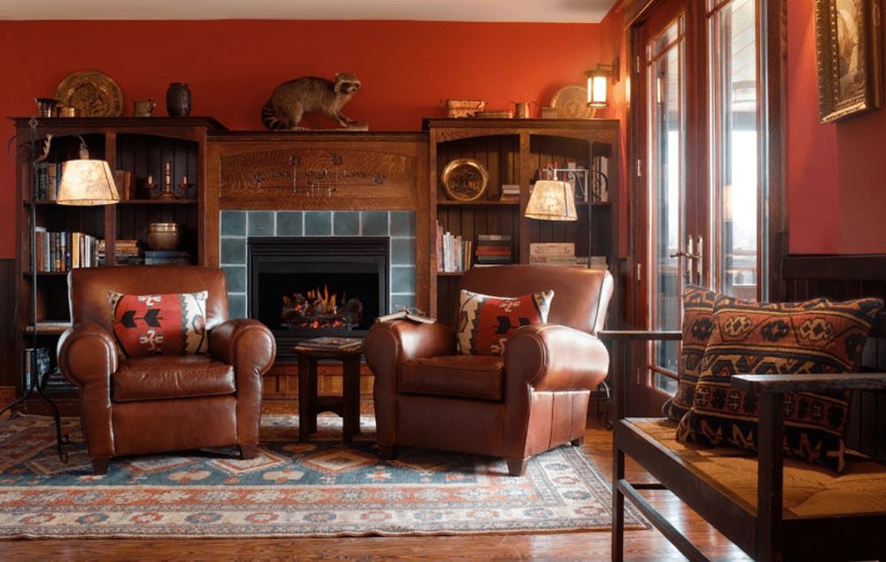 A pair of brown leather armchairs sit in front of the fireplace flanked by wooden shelvings. It has wrought iron floor lamps and a vintage rug that lays on the hardwood flooring.