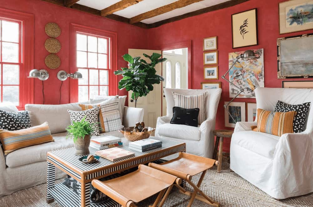 Fresh living room offers a fiddle leaf fig plant and white skirted seats paired with a striped coffee table. It has a gallery wall and a regular ceiling lined with rustic wood beams.