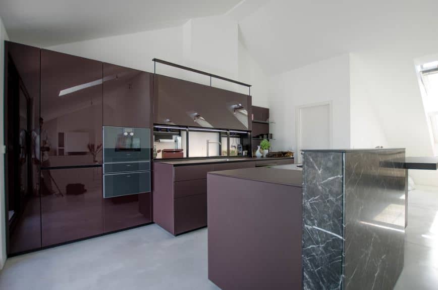 The white cathedral ceiling and gray concrete flooring are both contrasted by the large dark purple structures of the peninsula and kitchen island that has a raised breakfast bar attached to it made of green marble that goes well with the modern cabinets and drawers.