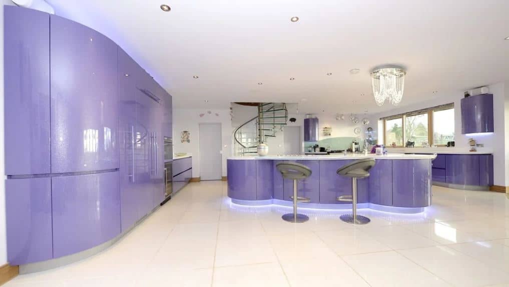 The sleek purple elements of this modern kitchen has a curved design to it that veers away from the traditional lines and edges. This is augmented by the lights underneath the structures that casts blue lights onto the white flooring surrounding the kitchen island.