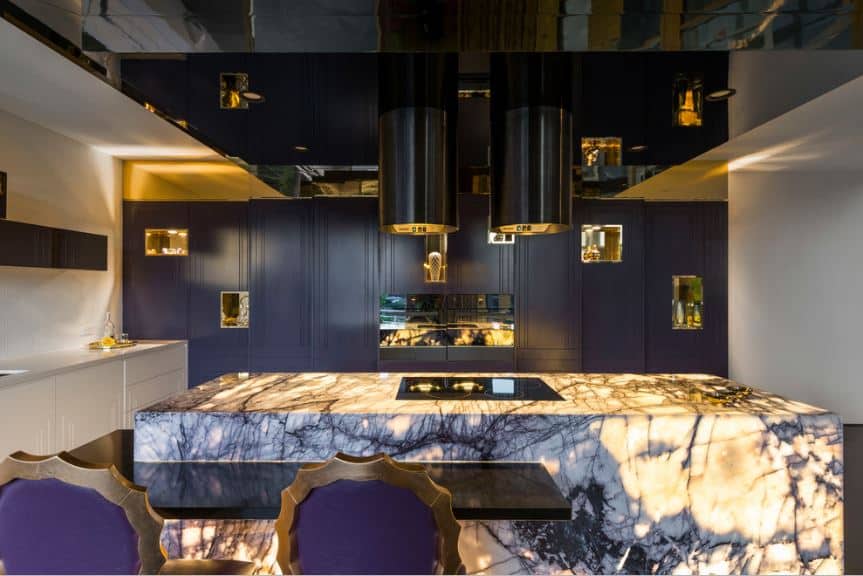 This eclectic and modern kitchen has a waterfall kitchen island made of white and purple marble that sets an amazing foreground for the rest of the kitchen that has dark purple cabinetry complemented by the slick black ceiling panels.