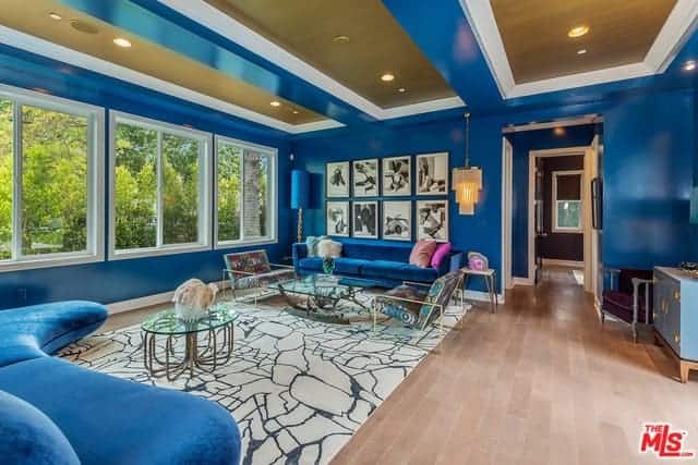 This living room is designed with a photo gallery mounted above the blue velvet sofa that's lighted by a cascading chandelier. It is accompanied by modern seats and glass top coffee tables on a brown area rug over the wide plank flooring.