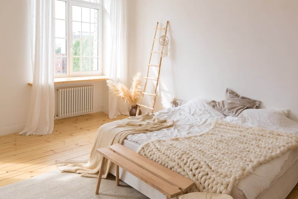 A focused shot at this primary bedroom's large bed set on the hardwood flooring. The glass window on the side features lovely white window curtains.