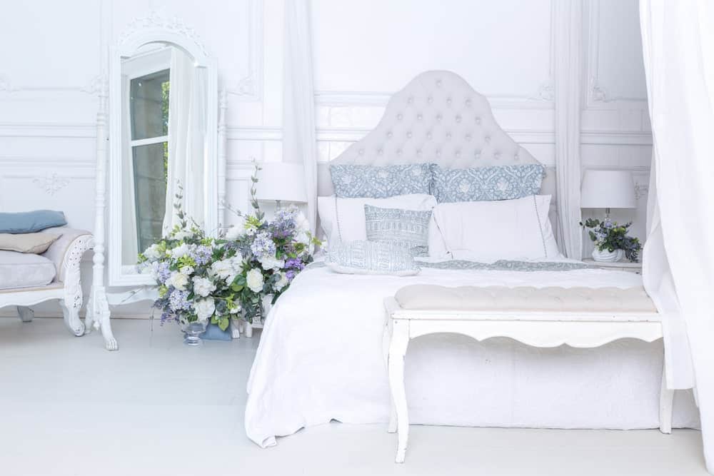 Large primary bedroom featuring a gorgeous bed setup with beautiful flowers on its side lighted by table lamps and is surrounded by white walls and floors.
