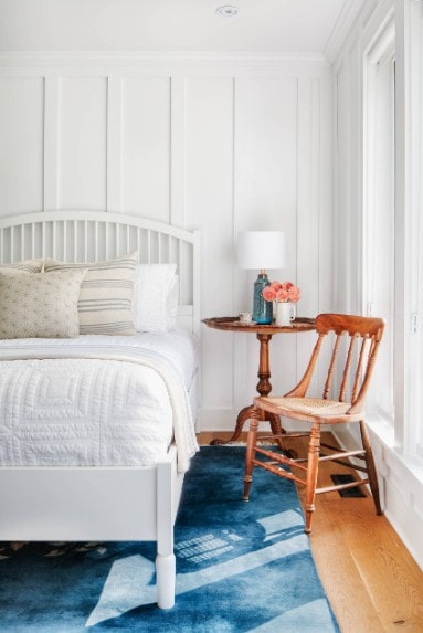 This primary bedroom offers a large white bed matching the white walls and white ceiling. The hardwood flooring is topped by a stylish blue area rug.