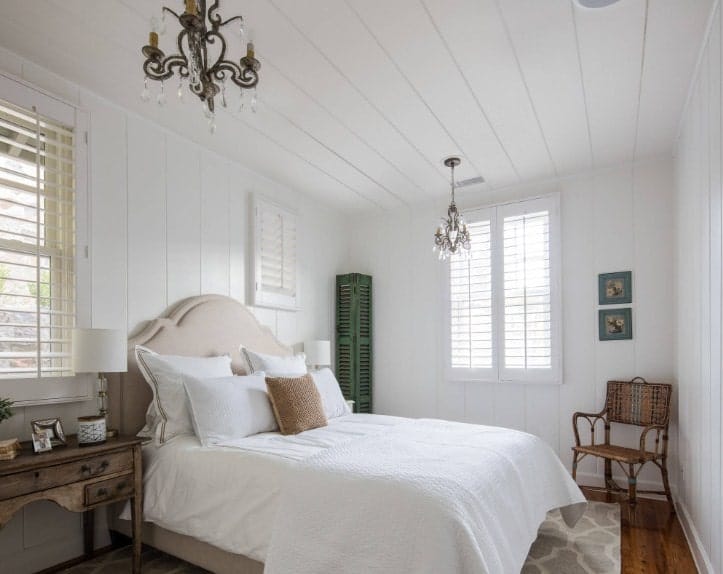 This primary bedroom features a classy bed setup lighted by two gorgeous chandeliers and is surrounded by white wooden walls and ceiling.