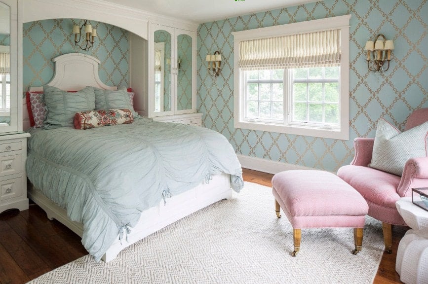 Primary bedroom with elegant green walls lighted by wall lights. The room offers a large classy bed along with a pink chair with a footrest on the side. The hardwood flooring is topped by a white area rug.