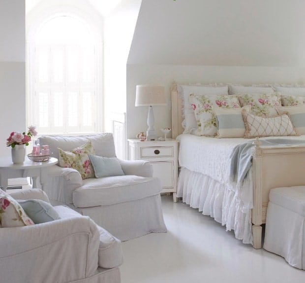 White primary bedroom featuring white flooring and white walls. The room offers a large lovely bed along with two white seats on the side with a small center table.