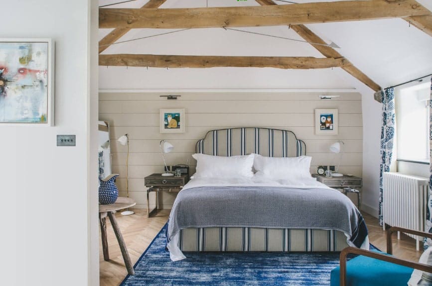 Large primary bedroom featuring a classy bed set on top of a stylish blue area rug covering the herringbone-style hardwood flooring.