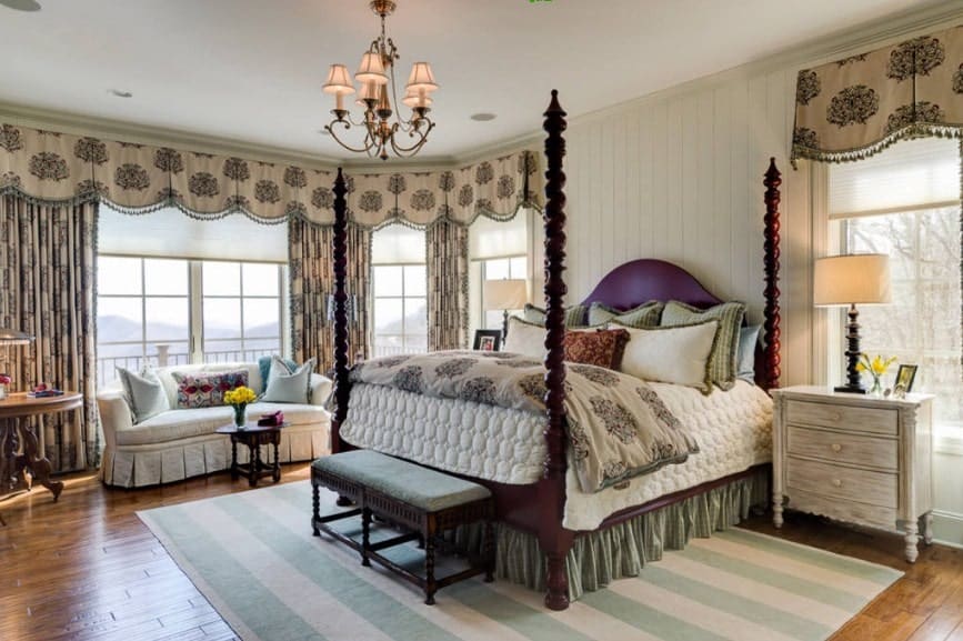 Large primary bedroom with elegant window curtains and a gorgeous chandelier. The room has a large cozy bed and a nice couch near the windows.