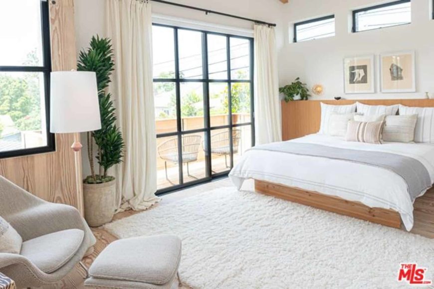 This is an airy and bright Scandinavian-style bedroom with its white ceiling and walls matched with the brightness from the French sliding glass doors leading to a balcony. The traditional wooden bed melds with the hardwood flooring that is covered by a white area rug.