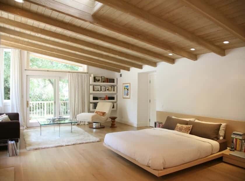 Large Scandinavian-Style primary bedroom featuring a glamorous wooden ceiling with beams matching the hardwood flooring. The bed looks very stylish and romantic. There’s a reading nook on the side as well.