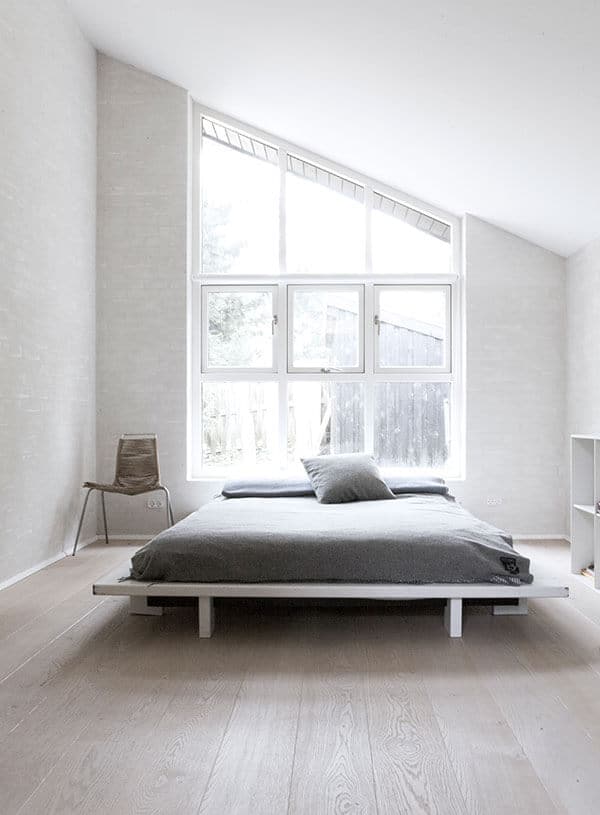 The white shed ceiling of this Scandinavian-Style bedroom is complemented by a tall window that follows the shape of the ceiling. It is at the head of the bed cushion that is on a charming white wooden platform that matches the white floor and walls.