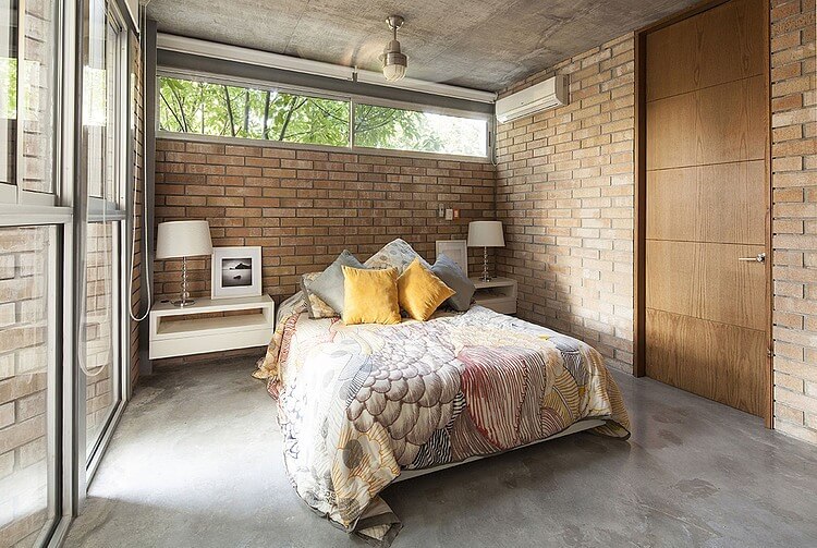 The gray concrete flooring of this industrial-style bedroom matches with the gray concrete ceiling that supports a semi-flush mounted light over the bed with a colorful sheet that complements the red brick walls.