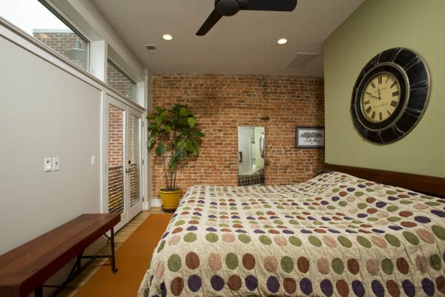 The colorful patterned sheet of the bed is paired with a low wooden headboard that is topped with an antique bulky wall-mounted clock on a light green wall. This is contrasted by the adjacent red brick wall with a potted plant in the corner.