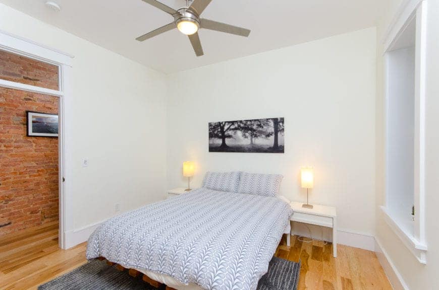 This bedroom has a stainless steel industrial-style ceiling fan mounted on the white ceiling that matches the white walls and contrasted by the hardwood flooring as well as the red brick wall just outside the door.
