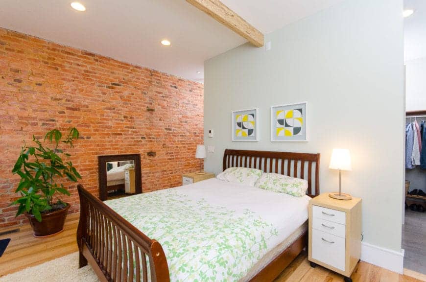 The wooden sleigh bed has a wooden headboard with a slat finish that stands out against the light gray wall topped with a white ceiling with a single wooden beam in the middle. This pairs well with the red brick wall and hardwood flooring.