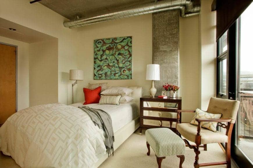This charming primary bedroom is a lovely fusion of traditional-style and industrial-style. It has a traditional bed that complements the beige carpeted flooring and beige walls. This is accented with exposed pipes and vents of the concrete ceiling.