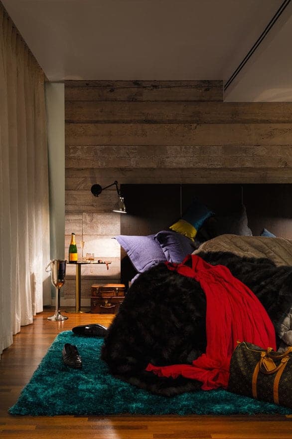 The black cushioned headboard of the traditional bed is against a wall made of distressed wooden planks that stand out against the white ceiling and the redwood flooring. This wall supports a wall-mounted lamp over the stainless steel bedside table.