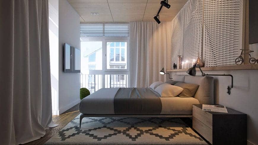 The industrial-style ceiling supports a pair of black spot lights that illuminated the light gray bed augmented by a couple of industrial-style wall lamps over the wooden bedside drawers over a patterned area rug.