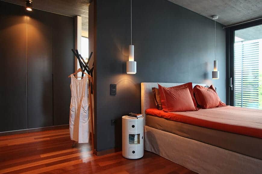 The gray concrete ceiling hangs a couple of modern white pendant lights on either side of the bed. these lights stand out against the black wall of the bed together with the white barrel-like bedside tables that contrast the redwood flooring.
